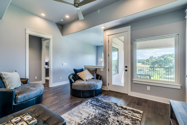 A modern living room featuring a round, cushioned chair, a sleek leather armchair, and a patterned rug. Large windows open to a scenic balcony view of lush greenery. The room has stylish decor with recessed lighting, a ceiling fan, and warm hardwood flooring, showcasing contemporary interior design ideas.